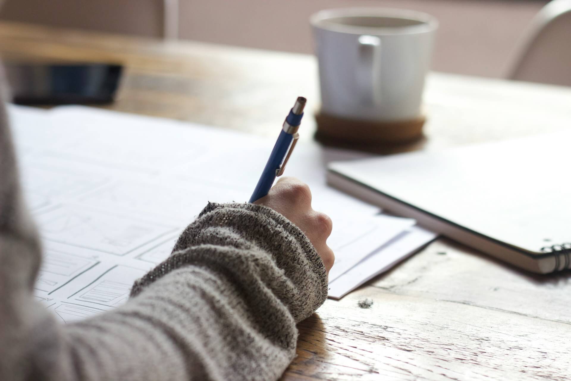 Person in a gray jumper holding a pen over paperwork at a desk with a coffee mug and notebook